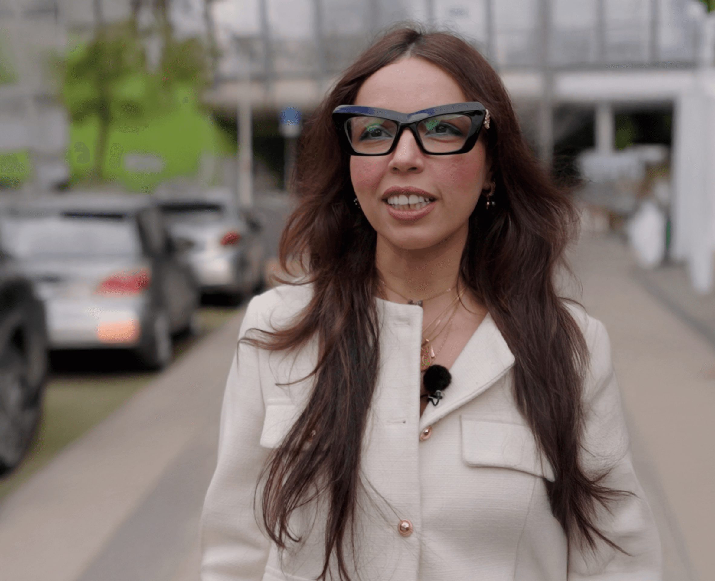 Nourhan staat buiten op een stoep langs geparkeerde auto's en kijkt lachend de camera in.