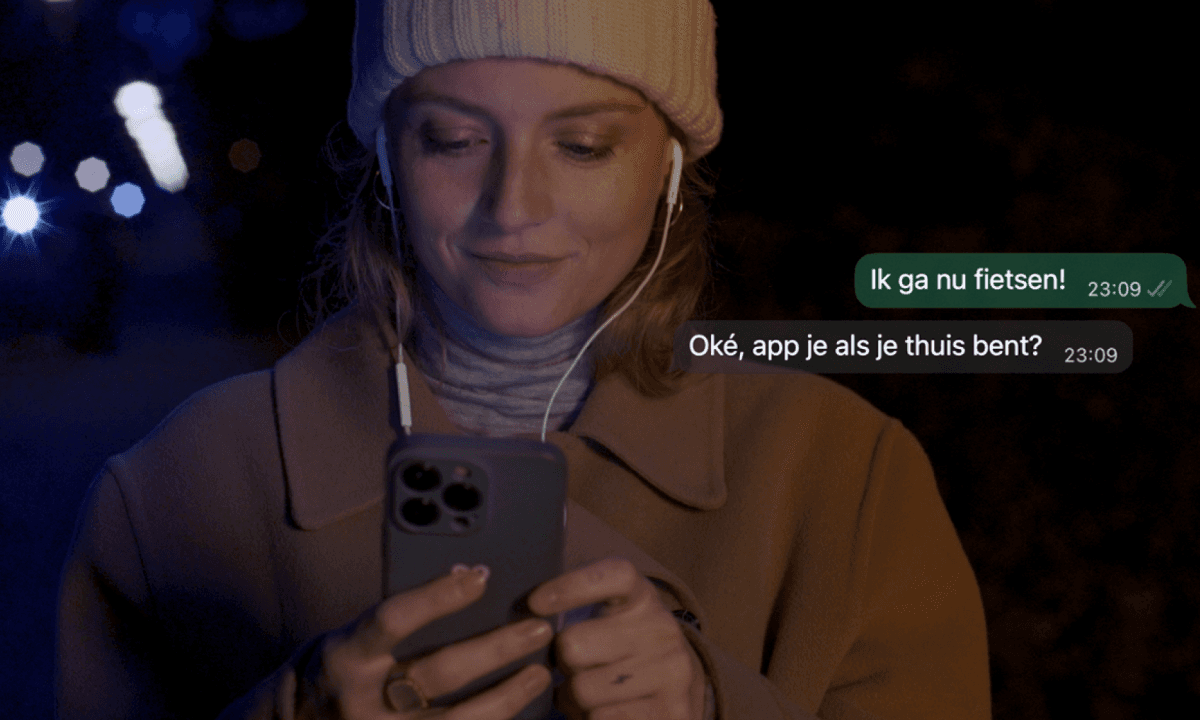 Een vrouw met een muts en oordopjes kijkt 's avonds lachend naar haar telefoon. Naast haar staan de berichten 'Ik ga nu fietsen!' en 'Oké, app je als je thuis bent?'.