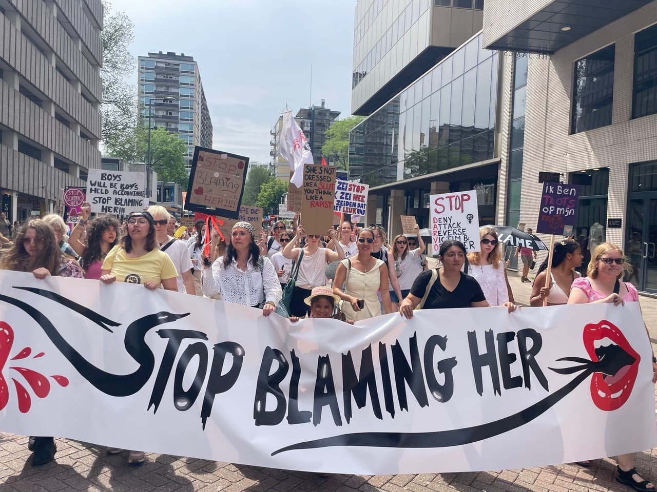 Een groep vrouwen loopt door een straat achter een breed spandoek met de tekst Stop blaming her, met protestborden erboven.